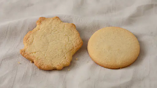 One uneven hand-cut uniced sugar cookie with an irregular wavy edge rests beside one smooth round-edged cookie on a plain cloth in gentle light.