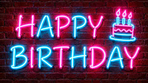 The words HAPPY above BIRTHDAY in glowing alternating hot pink and cyan neon tubing on a dark red brick wall with a small neon birthday cake with three candles beside them and a pink and blue glow on the bricks.