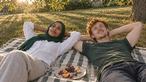 A woman in an emerald-green hijab and a slim man with tousled copper-red hair lie back side by side on a striped picnic blanket with their hands behind their heads beside a half-eaten plate of fruit and two empty tumblers with long shadows across the lawn.