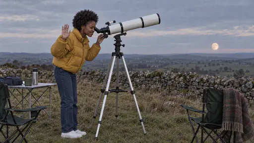 A young woman in a mustard puffer jacket stands with one eye at the eyepiece of a white refractor telescope and a hand on the focus knob aimed at a pale moon low over a drystone wall in faint twilight.