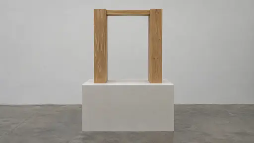 Two joined upright wooden pieces frame one plain rectangular opening on a pale plinth against a plain wall in even gallery light.
