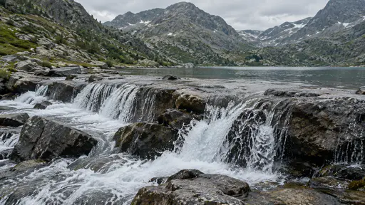 A lake outlet crosses a low rock sill at the end of a mountain lake with the water sheeting over the stone before continuing below.