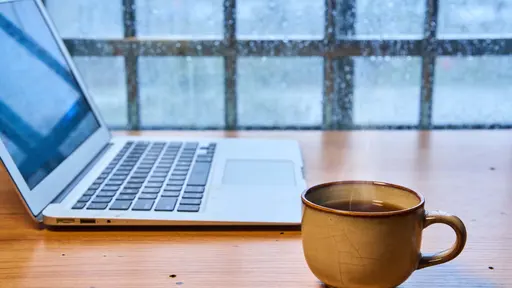Cozy 16:9 lifestyle photo of a laptop and steaming coffee on a wooden desk beside a rain-speckled window, rendered with soft diffuse light and moody atmosphere.