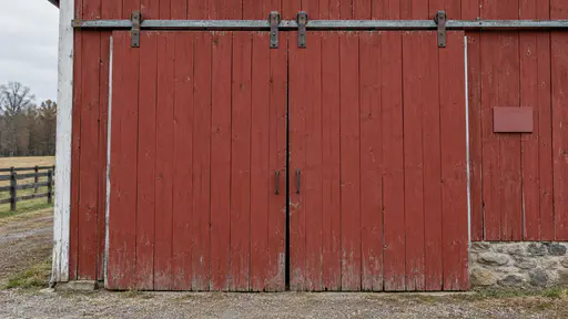 The large sliding door of a red barn fills the frame with its upper track and plain rollers visible above the door panels in soft side light.