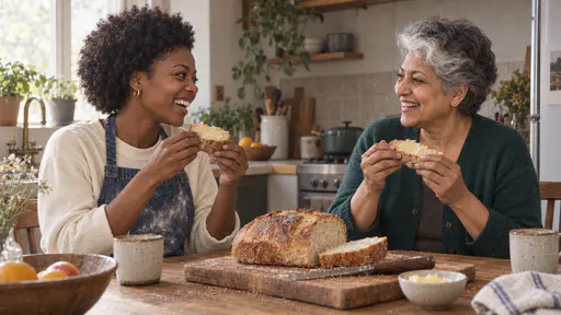 A young woman with a short natural afro in a flour-dusted denim apron and a South Asian woman with short silver-grey hair and a white streak in a green cardigan laugh warmly across a sunlit kitchen table each holding a thick buttered slice of fresh golden bread.