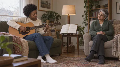 A young woman with a short natural afro concentrates on a plain acoustic guitar alone on a sofa while a South Asian woman with short silver-grey hair and a white streak watches with calm pride from an armchair with her hands folded in her lap in quiet late light.