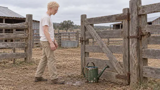 A slim man with albinism in his late twenties sets down a filled battered green enamel watering can with copper staple stitches just inside a weathered corral gate and steps back with empty hands in soft ranch morning light.