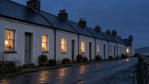 A long terrace of identical small cottages at deep blue dusk shows warm lit windows along most of the row, the farther windows dimmer, and a single lamp lit in the last cottage's window at the end.