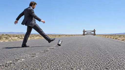 Kicking the can down the road: a suited adult kicks a lone dented steel can down an empty asphalt road toward a small unfinished timber barricade on the horizon.