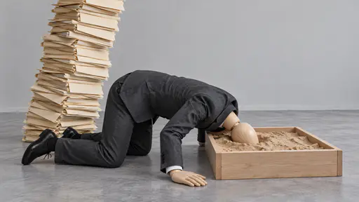 Burying your head in the sand: a suited mannequin kneels with its head hidden inside a sandbox on an office floor while a leaning tower of blank folders looms behind.