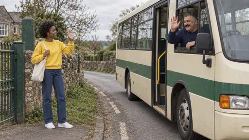 A heavyset bus driver with a wide moustache leans from the driver's window of a small cream and green rural bus with a raised hand while a young woman with a short natural afro and a canvas bag waves back from the verge at a school gate in bright light.