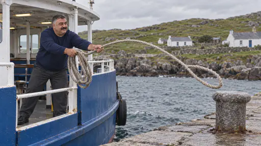 A heavyset deckhand with a wide moustache in a navy sweater leans out from the side of a small blue and white ferry and throws a coiled mooring rope over a bollard on a grey stone island jetty in clear midday light.