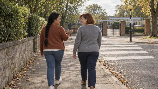 A woman with a long black braid in a rust cardigan gestures ahead with an open hand toward a zebra crossing while a woman with an auburn bob in a grey sweater listens and nods as they walk a tree-lined street toward a distant plain school gate.