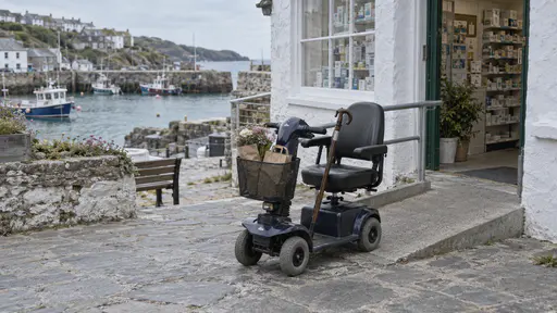 A mobility scooter with a front basket holding a paper bag and a bunch of flowers is parked outside a small harbour shop beside a ramp, its seat empty and a walking stick hooked on the handlebar in soft morning light.