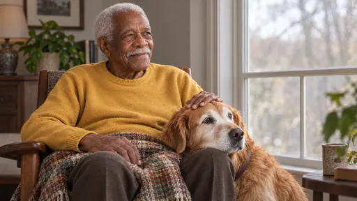 An elderly man with close-cropped white hair and a white moustache sits in a wooden armchair with a folded blanket over his knees, his hand resting on the head of an old golden retriever with a grey-white muzzle laid on his knee, both looking toward a window in soft light.