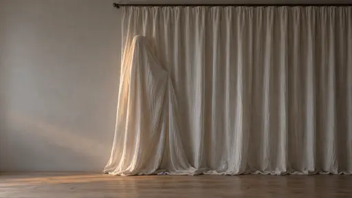A pale floor-length curtain hangs in a plain dim room; one fold near its far edge rises into the rounded silhouette of a sheet ghost with a wavy trailing hem, catching soft diffuse window light. A subtle haunted Halloween background image with clear empty copy space on the left.
