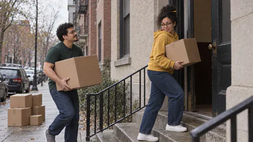 A tall man with round glasses and a chin beard in a green t-shirt carries a large cardboard box up the steps of a block of flats while a woman with round tortoiseshell glasses and a curly black bun in a mustard hoodie holds the heavy door open with her back and carries a smaller box.