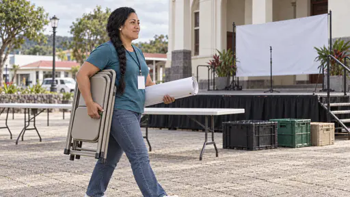 A Samoan woman with a long black braid and pearl studs in a teal shirt and jeans carries a stack of folding chairs under one arm and a rolled blank banner under the other across a small town square toward a small stage in front of a community hall on a bright morning.