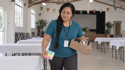 A Samoan woman with a long black braid and pearl studs in a teal shirt and lanyard carries a box of small tools and a stack of blank colour-banded tickets past a row of trestle tables with plain paper tablecloths in a bright community hall with bunting on the beams.