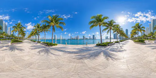 A full 360 degree equirectangular panorama from a paved plaza on a palm lined waterfront with a calm blue bay and a high rise glass skyline along the shore under a clear sky.