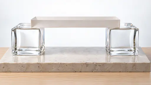A folded white paper bridge deck with low side walls rests across the tops of two square clear glass block piers on a pale stone plinth with a pale wooden base, held up a hand's height above the plinth in bright backlight.