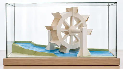 A large round paper wheel with eight flat paddles stands on a thick paper axle between two paper posts, its lowest paddles dipping into a thin blue paper stream inside a clear rectangular glass display case on a pale wooden base.
