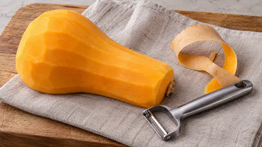 A whole peeled butternut squash lies on pale linen over an oak board showing smooth bright orange flesh, a long curl of tan peel and a steel peeler on the linen beside it.