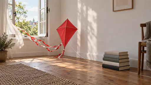 Perfectionism keeps the kite indoors — a flawless finished red kite stands ready on a wooden floor, its tail streaming in the breeze of a wide open window, but its string is pinned beneath a heavy stack of hardcover books, picturing the meaning of perfectionism: endless refinement preventing a finished work from being released.