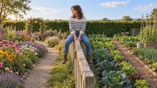 Sitting on the fence: a frontal full-body editorial garden photograph in soft afternoon light, where a person sits balanced astride the top rail of a narrow wooden fence on the boundary between a flower-filled garden path on the left and a vegetable-plot path on the right, leaning neither way.