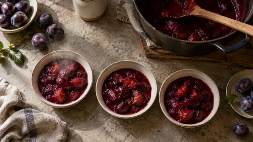 From directly above in gentle overhead light shallow white dishes of freshly cooked purple plum compote cool in a row beside a heavy preserving pan with a wooden spoon across it with steam still rising faintly from the nearest dish and a few whole plums beside.