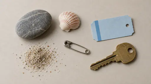 A flat-lay on a pale driftwood-coloured surface of one smooth grey pebble, a small pink shell, a blank blue paper ticket stub with a punched hole, a safety pin, a pinch of fine sand and a brass door key.