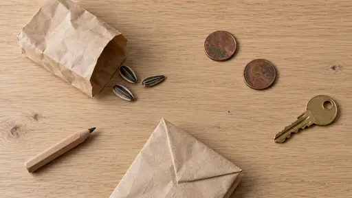 A flat-lay on a pale oak table of two worn copper coins, a small tipped brown paper bag with three sunflower seeds spilled beside it, a short pencil stub and a single brass door key with generous empty space around them.