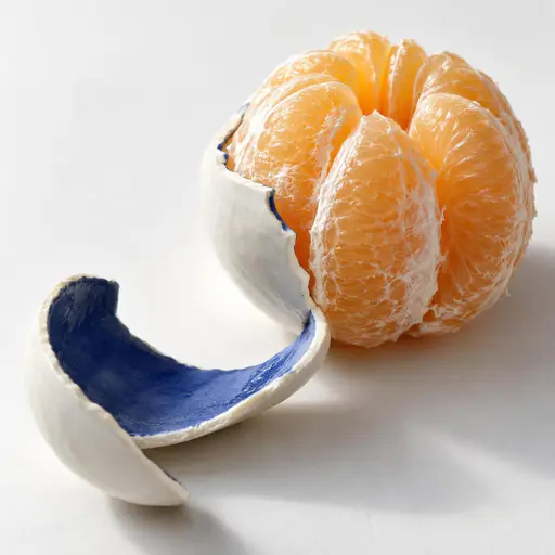 Close three-quarter view of a peeled citrus fruit with translucent segments on a chalk surface, one thin porcelain ribbon with a cobalt painted interior curling away, its jagged broken edge turned toward the glowing segments.