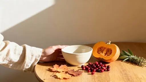 A warm, cinematic close-up of an early-morning kitchen table corner showing a matte cream ceramic bowl, halved sugar pumpkin, fresh cranberries, herbs, and scattered amber leaves.