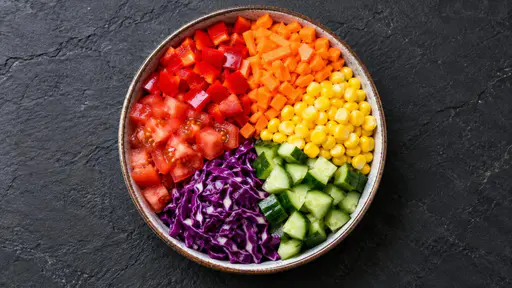A top-down view of a bowl filled with colorful chopped vegetables arranged in sections on dark slate.