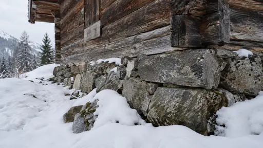 The raised stone footing of a cabin shows above uneven snowy ground with the timber wall starting above the stones in clear winter light.