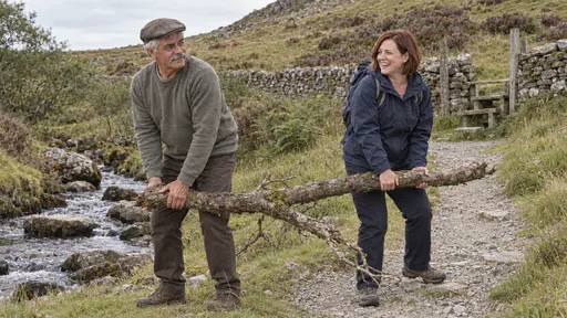 An elderly man with a grey moustache in a flat cap and a woman with a chin-length auburn bob in a navy jacket carry a fallen branch off a hill path beside a small stream with stepping stones, the woman laughing, in warm midday light.