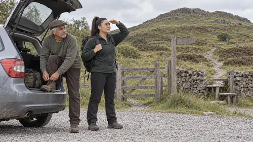 An elderly man with a grey moustache in a flat cap leans on the open boot of a small hatchback lacing his walking boot while a broad-shouldered woman with a rucksack and a high ponytail shades her eyes toward a green hill at a gravel trailhead.