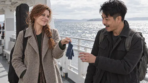 A freckled woman with a loose auburn braid holds out a small blue knitted glove between two fingers with a smile to a wiry man in a charcoal jacket who reaches for it with a surprised laugh on a ferry deck with a rail and a sparkling sea behind them in bright breezy light.
