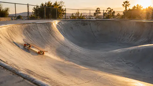 A skateboard with orange wheels rolling riderless down the curved wall of an empty concrete skatepark bowl at sunset, a trail of dust behind it, a chain-link fence and trees along the rim and low mountains in the distance.