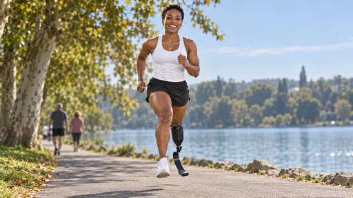 A lean woman with close-cropped hair and small silver hoops runs mid-stride on a paved lakeside path with a carbon-fibre blade on her right leg and a white shoe on her left foot, grinning, two slower walkers small and blurred behind her.