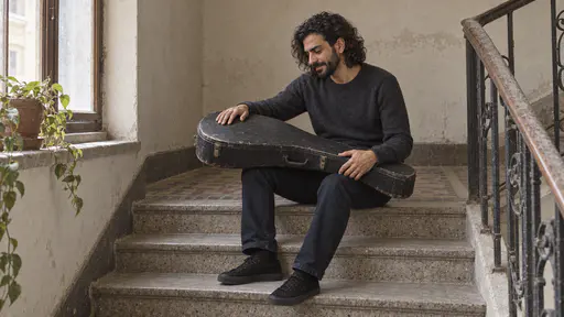 A slim bearded musician with thick wavy black hair sits on the top step of a stair landing with a worn black oud case across his knees and one hand laid flat on its lid with a fond expression in warm window light.