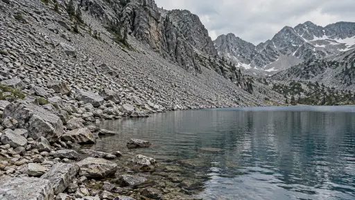 A grey scree slope of loose rock runs down to the water edge of a mountain lake with fallen stones along the waterline in clear daylight.