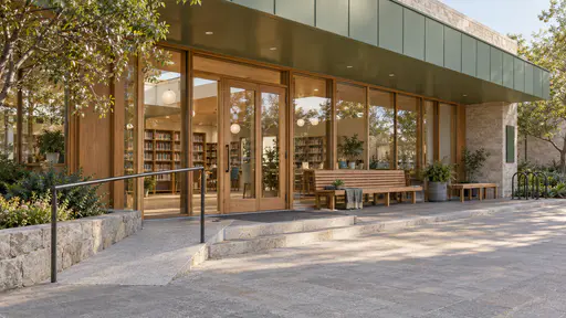 A long timber bench with a backrest and a shorter bench stand beside glass library doors under a sage green canopy with a folded blanket, a small potted plant and a bike rack at the far end in warm morning light.
