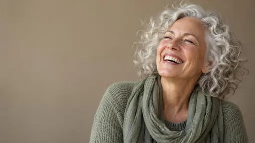 A woman in her sixties with silver curly hair laughs with her eyes closed, wearing a sage knit sweater and scarf against a plain warm taupe wall.