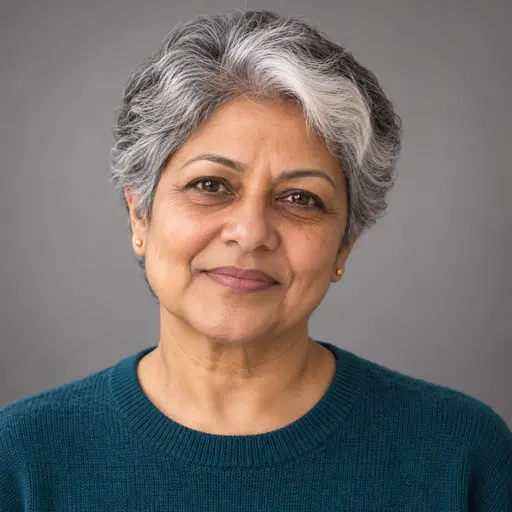 A close portrait of a woman in her mid fifties with short silver-grey hair cut to a sharp line, a wide white streak, small gold earrings and a deep teal sweater against a pale grey seamless backdrop, lit by a high frontal light.