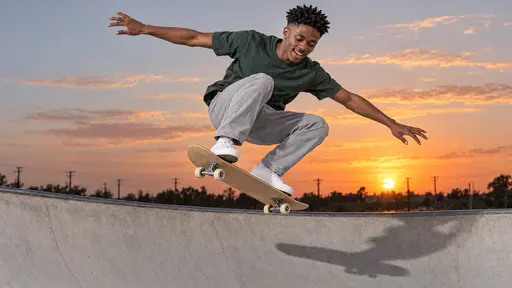 A young man with a coily twist-out in a forest-green t-shirt and white sneakers, airborne on a plain maple skateboard above the rim of a pale concrete skatepark bowl, arms spread and grinning, an orange sunset sky behind him.