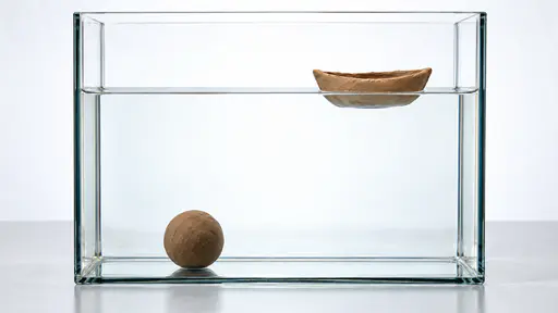 A solid clay ball lies sunk on the floor of a tall clear glass tank of water beside a hollow clay boat of the same clay floating upright at the waterline in front of a bright diffused backdrop.