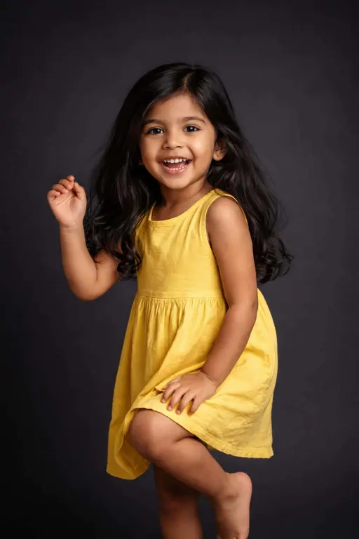 Cinematic 2:3 portrait of a four-year-old South Asian girl in a sunny-yellow dress, smiling toothily in a tiny dance pose against a deep charcoal backdrop with warm amber highlights and cool shadow edges.