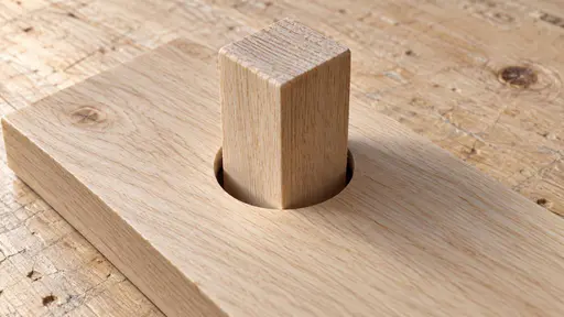 A square peg in a round hole: a high three-quarter macro product photograph under soft raking light, where a square wooden peg has been pressed partway into a perfectly circular opening in a pale timber board, its four corners resting on the round rim and holding it proud of the surface.